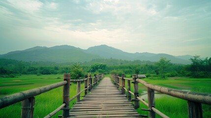 Obraz premium Serene wooden footbridge leading through lush green fields towards distant mountains under a cloudy sky, evoking a sense of calm and exploration.