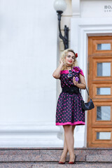 Blonde girl in a bright dress and sunglasses on the street near a building on a walk on a summer day. Hipster