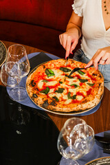 Cliente di una pizzeria elegante mentre taglia al tavolo un pizza margherita tradizionale con sugo di pomodoro, mozzarella e basilico fresco