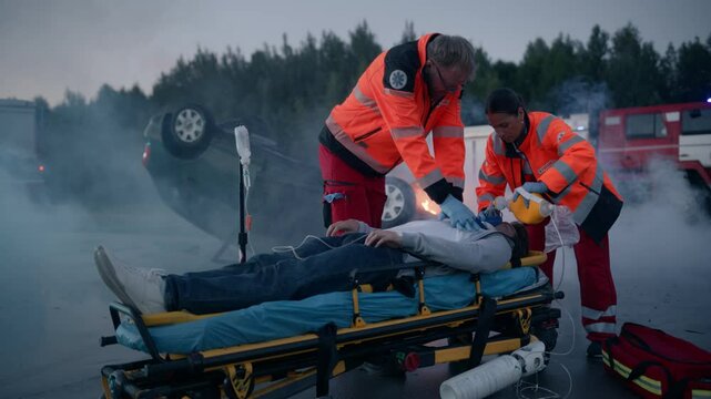 Ambulance Medics Doing Cardiopulmonary Resuscitation to an Unconscious Patient After a Serious Road Accident with a Rolled Car. Scene with Burning Car, Firefighters and Fire Engines in the Background