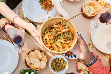 Hands passing a bowl of pasta at a table with various dishes, seen from above. Diverse group...