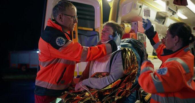 Medical Rescue Team Inspecting the Health of a Traffic Accident Survivor. Young Male Patient Lying on a Stretcher in an Ambulance, Doctor Using a Flashlight to Diagnose a Potential Brain Trauma