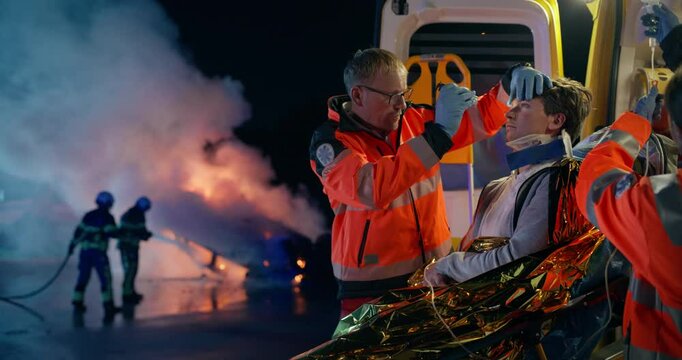Rescue Scene at Night with a Middle Aged Paramedic Diagnosing Injuries on a Young Patient with a Flashlight in an Ambulance while Firefighters Extinguish a Burning, Rolled Vehicle