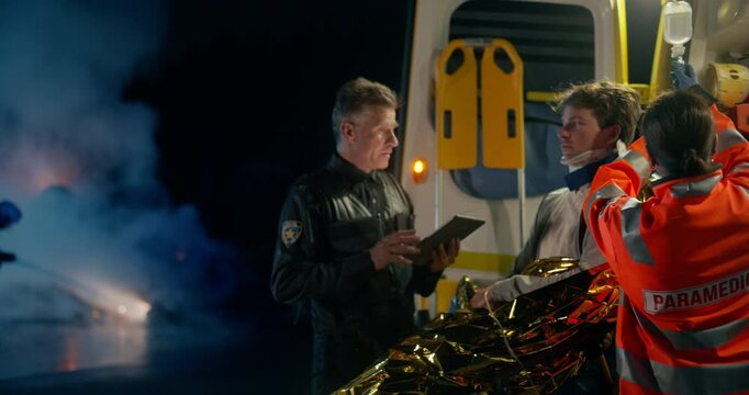 Policeman Using a Tablet Computer and Writing Down a Report From a Car Crash Victim. Law Enforcement Officer and a Paramedic Helping a Young Man on a Stretcher. Firemen Extinguish an Overturned Car
