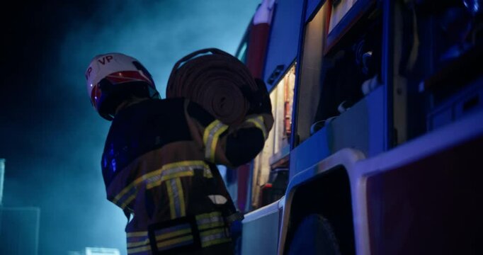 Cinematic Scene with a Firefighter Crew Preparing to Battle a Car Fire at a Traffic Accident at Night. Fireman Taking a Rolled Up Power Hose from the Fire Engine, Bringing it Closer to the Accident