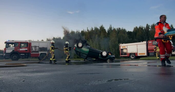 Emergency Responders Extinguish an Overturned Car Fire while Paramedics Assist an Injured Victim. Firefighters Use High Pressure Hoses to Control the Blaze. Car Accident Scene with Emergency Vehicles