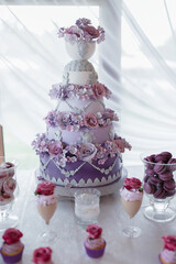 A tall, multi-layered cake with purple flowers and a white base. The cake is placed on a table with other desserts and drinks. Scene is celebratory and festive