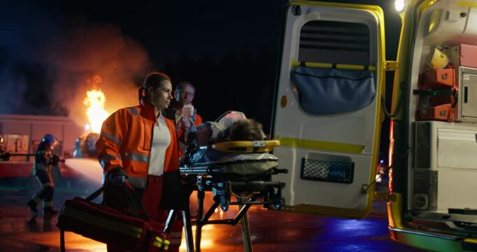 Dramatic Nighttime Rescue Unfolds as Firefighters Battle Car Fire while Paramedics Rush to Evacuate Injured on a Stretcher. Flames Illuminate a Coordinated Emergency Response In a Smoke-Filled Street