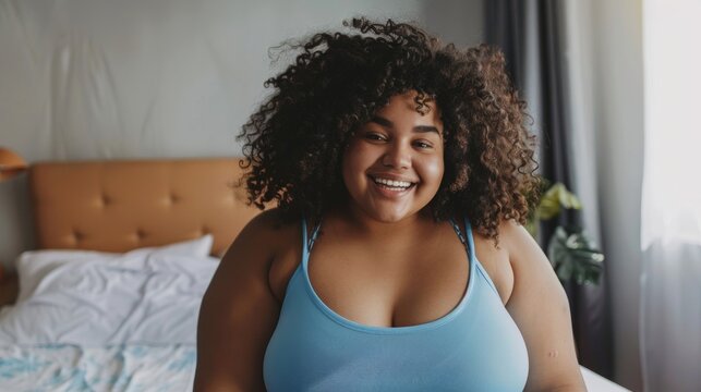 Smiling woman with natural hair, dark skin, perfect teeth, and curvy figure in light blue tank top sitting on bed in softly lit pastel bedroom.
