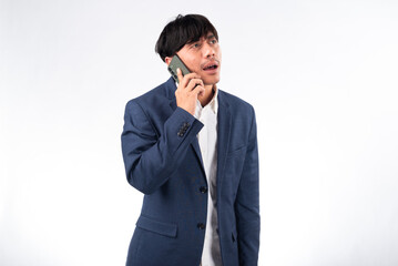 A surprised Asian businessman in a navy blue suit holds a smartphone to his ear, reacting with shock during a phone call. The white background emphasizes his expression