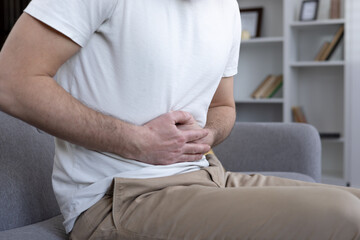 Obraz premium Man holding stomach in discomfort, indicating abdominal pain. Clad in casual attire, seated on sofa in modern living room. Captures concept of health issues, illness, or indigestion.