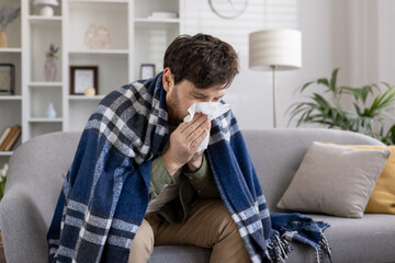 Man sitting on couch wrapped in a blue blanket, sneezing into tissue. Scene depicts illness, cold, flu symptoms, and seeking comfort at home. Expression of discomfort and need for care.