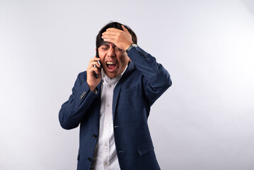 An Asian businessman in a navy blue suit displays extreme distress while holding a smartphone to his ear. His hand grips his head in frustration, emphasizing a tense phone call. White background
