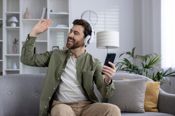 Smiling man listening to music on headphones while holding phone. Relaxing on couch, expressing joy and connection. Casual home setting captures leisure and happiness.