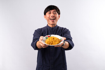 An enthusiastic Asian man in a dark shirt presents a plate of noodles with a big smile, showcasing a delicious meal. The plain white background emphasizes his inviting gesture.