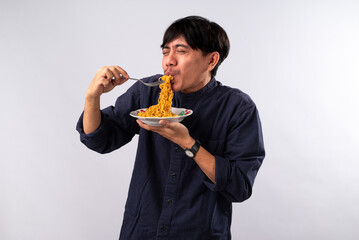 An Asian man in a dark shirt eagerly eats noodles from a plate, his expression filled with delight....