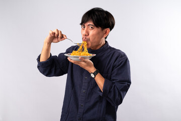 An Asian man in a dark shirt eagerly eats noodles from a plate, his expression filled with delight....