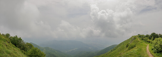 Fototapeta premium ampia vista panoramica dalla cima della dorsale erbosa di un alto monte nel nord est Italia, sotto un cielo nuvoloso e coperto da foschia, di giorno, in estate
