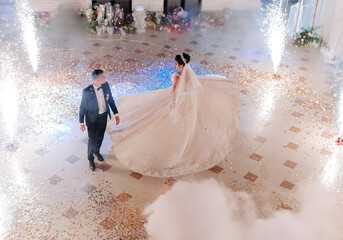 A bride and groom are walking down a dance floor with fireworks in the background. The bride is wearing a long white dress and the groom is wearing a suit. Scene is celebratory and romantic