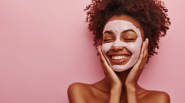 A happy woman with a facial mask, isolated on a pastel pink background. A beauty portrait of a smiling African American girl, smiling while washing her face during her morning skincare routine. 
