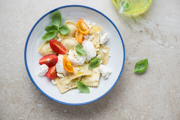 Italian ravioli served with ricotta, tomatoes and basil, flat lay on a beige stone background, horizontal shot