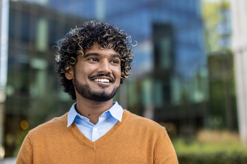 Confident Indian businessman smiling in urban environment, wearing orange sweater and blue shirt. Depicts professional success and positive outlook. Concepts of business confidence, optimism