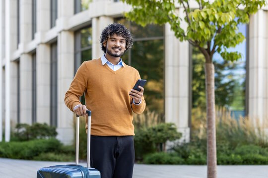 Smiling man stands outdoors with phone and suitcase, conveying readiness and confidence. He is dressed casually, suggesting a blend of business travel and leisure. - Powered by Adobe
