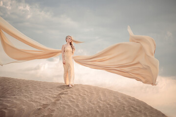 A blonde girl in a beautiful beige dress stands on a dune in the desert. the dress flutters...