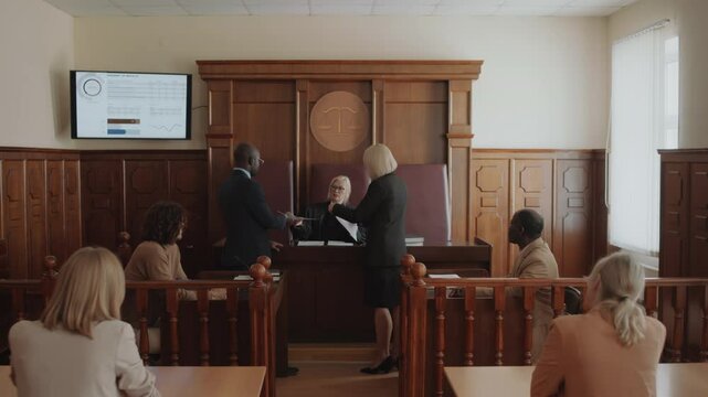 Female judge handing out juridical documents to male attorney and female prosecutor standing in front of judges bench, then they taking seats in courtroom