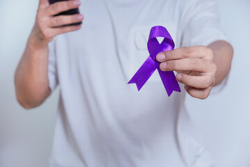 A person holds a purple ribbon in one hand while using a smartphone, symbolizing awareness and...
