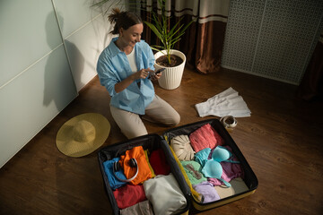Woman organizes clothes and a sun hat while checking a packing list on her smartphone. Carefully...