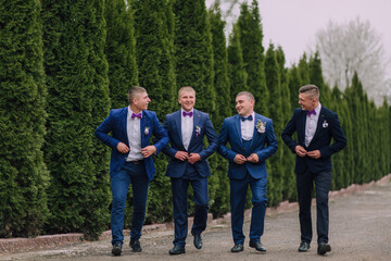 Four men in suits are walking together, with one of them wearing a purple bow tie. Scene is lighthearted and fun, as the men are dressed up and enjoying each other's company