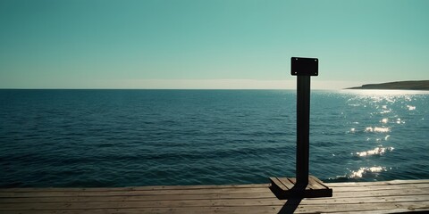 Fototapeta premium A weathered metal plate mounted on a pole at the end of a wooden quay bridge, overlooking the calm sea under a clear sky.