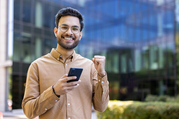 Smiling young man holding smartphone showing excitement and happiness in outdoor urban setting....
