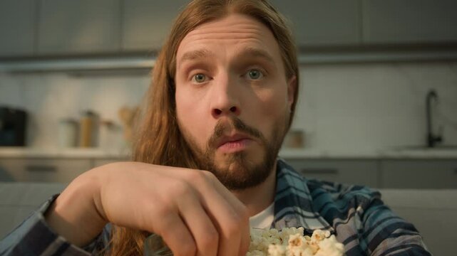Portrait concentrated focused funny Caucasian bearded man guy male at home couch sofa relaxing watching TV television series move film program sport championship football game eating popcorn obesity