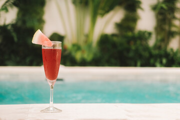 Banner with a glass goblet of red cold watermelon juice, which stands against the background of a pool with blue water and tropical green bushes. Juice detox. Cooling down in the heat. Background.