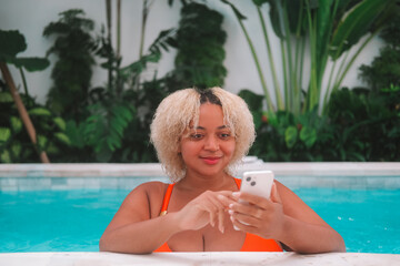 Young African American curvy woman in orange bikini relaxing in the villa pool and scrolling social media on her mobile phone. Using cell phone during vacation. Digital detox. Modern technologies.