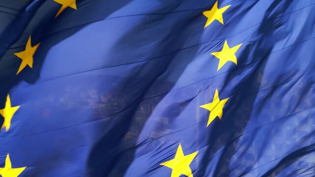 A close-up shot of the European Union flag waving in the wind, with its iconic blue background and golden stars. The movement of the fabric adds a dynamic and patriotic feel