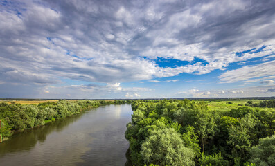 A tranquil river meanders through vibrant greenery, reflecting a stunning sky filled with clouds, creating a peaceful atmosphere in the early afternoon light.