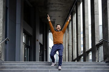 Joyful man celebrating success with raised arms on urban staircase outside modern building....