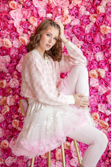 Beautiful girl in pink and white checkered jacket and white skirt posing in studio. She stands in front of pink wall with flowers.