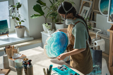 Craftswoman in respirator inspects dried epoxy resin painting, evaluating results in workshop....