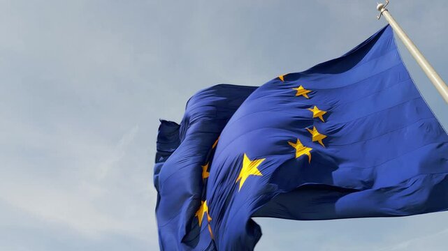 A close-up shot of the European Union flag waving in the wind, with its iconic blue background and golden stars. The movement of the fabric adds a dynamic and patriotic feel