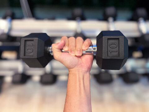 hand gripping dumbbell with a strong, determined hold. The background features a blurred view of other gym equipment, highlighting the focus on the dumbbell and the intensity of the workout, fitness