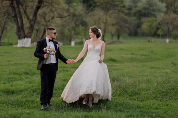 A bride and groom are walking together in a field. The bride is wearing a white dress and the groom is wearing a suit. They are holding hands and appear to be happy and in love