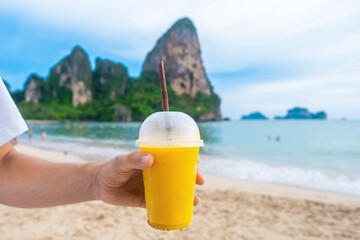 Man holding yellow mango smoothie on tropical sandy Railay beach with cliffs in the background at Krabi province, Thailand. Male hand holds refreshment summer drink in plastic cup with a straw