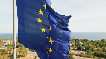 The European Union flag waves prominently above a coastal city, with the sea and urban landscape in the background. The scene blends patriotism with the beauty of nature and modern architecture - Powered by Adobe