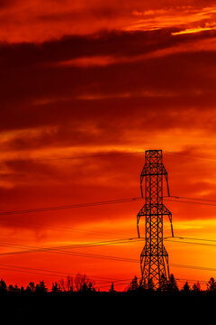 Silhouette of large, metal electrical transmission tower with a colorful, fiery red sky creating a dramatic background at twilight; Calgary, Alberta, Canada