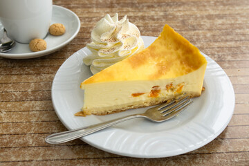On the table in a café, there is a plate with a piece of cheesecake and whipped cream. A cup of coffee is in the background. German cake quark-mandarin