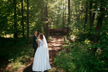 A bride and groom are standing in a forest, surrounded by trees. The bride is wearing a white dress and the groom is wearing a suit. They are both smiling and looking at the camera